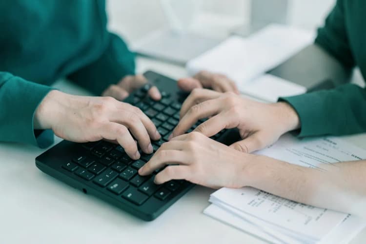 Mãos digitando em teclado de computador com documentos legais ao lado, foco no teclado.