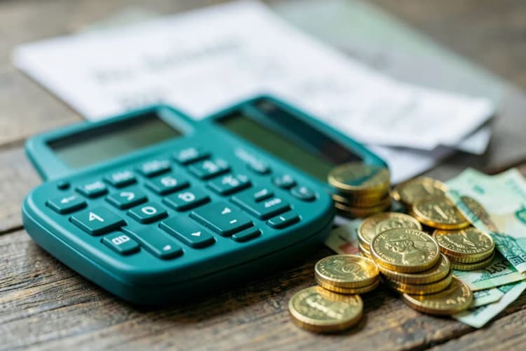 Uma calculadora e um monte de moedas de ouro e notas de dinheiro sobre uma mesa, com um contrato desfocado ao fundo.