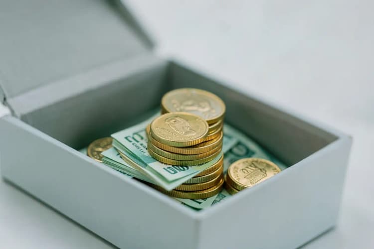 Uma pilha de moedas de ouro e notas de dinheiro sendo devolvidas de uma caixa aberta.